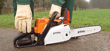 : Step 7 of 15: Leaving the chainsaw on the ground, place your left hand on the handlebar and your right hand on the starter rope. Push your right toecap into the rear handle to stabilize the chainsaw.